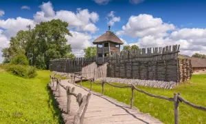 Drewniana kładka prowadzi do zrekonstruowanej osady Biskupin. Widać palisadę, wieżę strażniczą i chaty kryte strzechą.