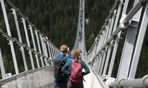 Para spaceruje po wiszącym moście w Czechach, robiąc zdjęcia. W tle widać las i innych turystów.