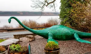 Zielony potwór z Loch Ness leży na kamieniach, z długą szyją wyciągniętą w stronę jeziora.