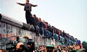 Tłum ludzi siedzi na Murze Berlińskim, świętując jego upadek. Jeden mężczyzna stoi na szczycie, wskazując palcem w dal.