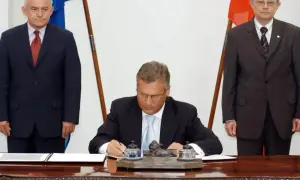 Uroczyste podpisanie dokumentów, kiedy Polska weszła do Unii. Trzech mężczyzn w garniturach, dwa flagi.