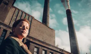Kobieta na tle dymiących kominów huty Szopienice. Ołowica unosi się w powietrzu, tworząc ponury krajobraz.