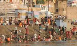Tłum ludzi na schodach nad rzeką Ganges, niektórzy kąpią się, inni siedzą i obserwują.