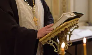 Duchowny w szatach, z krzyżem na szyi, czyta księgę podczas nabożeństwa prawosławnego.