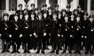 Grupa mężczyzn w czarnych mundurach i czapkach z orłem. Wśród nich widać symbole nawiązujące do gestapo.