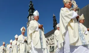 Biskup w białej szacie i mitrze z Matką Boską, na tle wieży kościelnej.