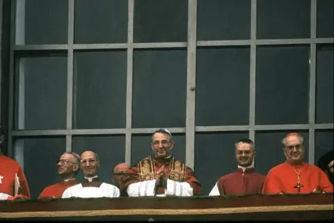 Kardynałowie w czerwonych szatach i jeden w białej sutannie, z papieżem w centrum. W tle kratowane okno.