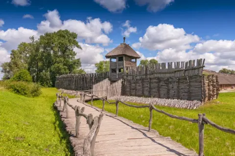 Drewniana kładka prowadzi do zrekonstruowanej osady Biskupin. Widać palisadę, wieżę strażniczą i chaty kryte strzechą.