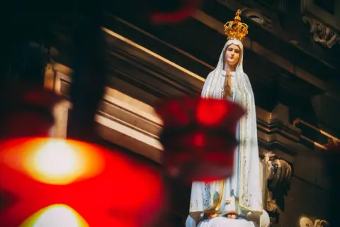 Figurka Matki Bożej Fatimskiej w koronie, z rękami złożonymi do modlitwy. W tle widać architekturę kościoła, a na pierwszym planie rozmyte czerwone światła, symbolizujące trzecia tajemnica fatimska.