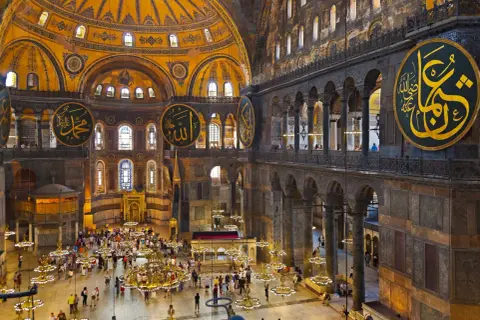 Wnętrze Hagia Sophia z wielkimi okrągłymi kaligrafiami, kopułą i tłumem turystów.
