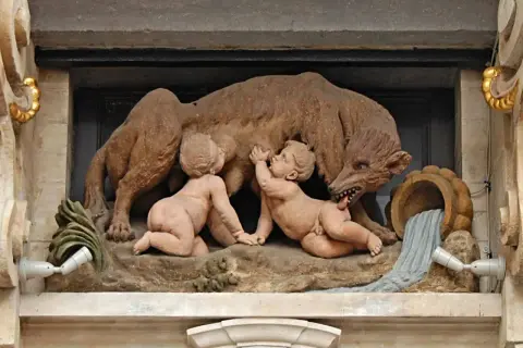 Rzeźba przedstawia wilczycę karmiącą Romulusa i Remusa, symbolizującą legendę o założeniu Rzymu. To klasyczny przykład motywu salut rzymski.