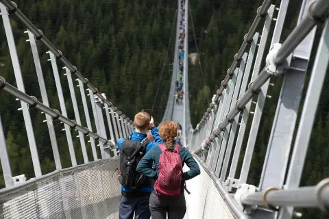 Para spaceruje po wiszącym moście w Czechach, robiąc zdjęcia. W tle widać las i innych turystów.