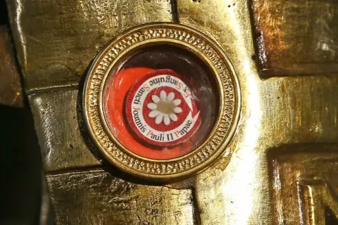 Złota oprawa zawiera relikwie, okrągły medalik z napisem "Ex Sanguine Sancti Joannis Pauli II Papae" i białym kwiatkiem.