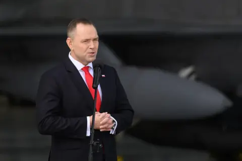 Mężczyzna w garniturze przemawia przy mikrofonie, w tle samolot. Mówi o reparacje wojenne od niemiec.