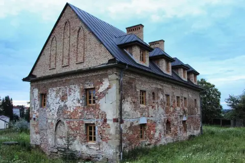 Zniszczony ceglany budynek z drewnianymi oknami i ciemnym dachem, otoczony zielenią.
