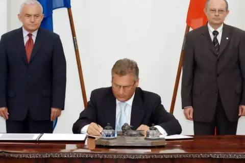 Uroczyste podpisanie dokumentów, kiedy Polska weszła do Unii. Trzech mężczyzn w garniturach, dwa flagi.