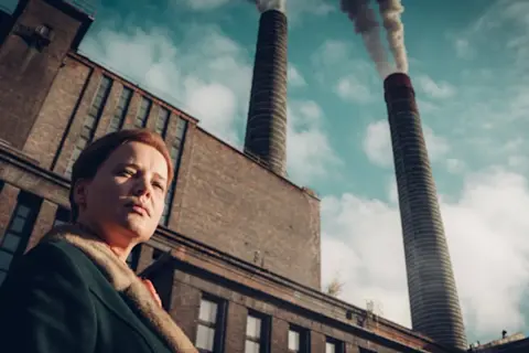 Kobieta na tle dymiących kominów huty Szopienice. Ołowica unosi się w powietrzu, tworząc ponury krajobraz.