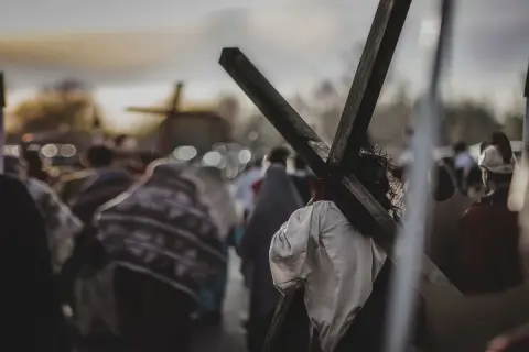 Osoba z koroną cierniową niesie krzyż, idąc w procesji. W tle widać inne krzyże i ludzi, przypominające stacje drogi krzyżowej.