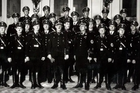 Grupa mężczyzn w czarnych mundurach i czapkach z orłem. Wśród nich widać symbole nawiązujące do gestapo.
