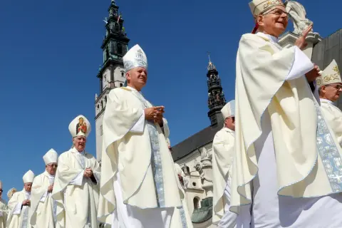 Biskup w białej szacie i mitrze z Matką Boską, na tle wieży kościelnej.