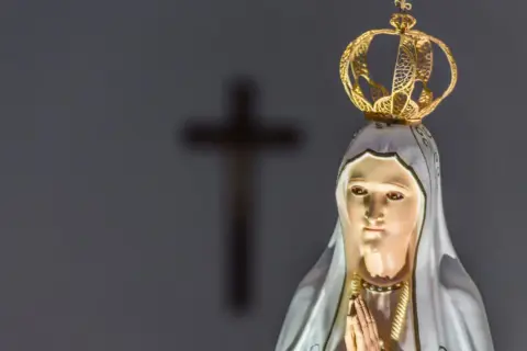 Figurka Matki Bożej z Fatimy, w koronie, z modlącymi się dłońmi. W tle niewyraźny krzyż, symbol objawienia fatimskiego.