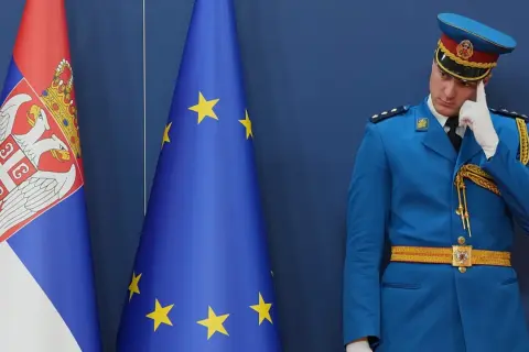 Strażnik w niebieskim mundurze zastanawia się, czy Serbia jest w Unii, stojąc obok flag Serbii i UE.