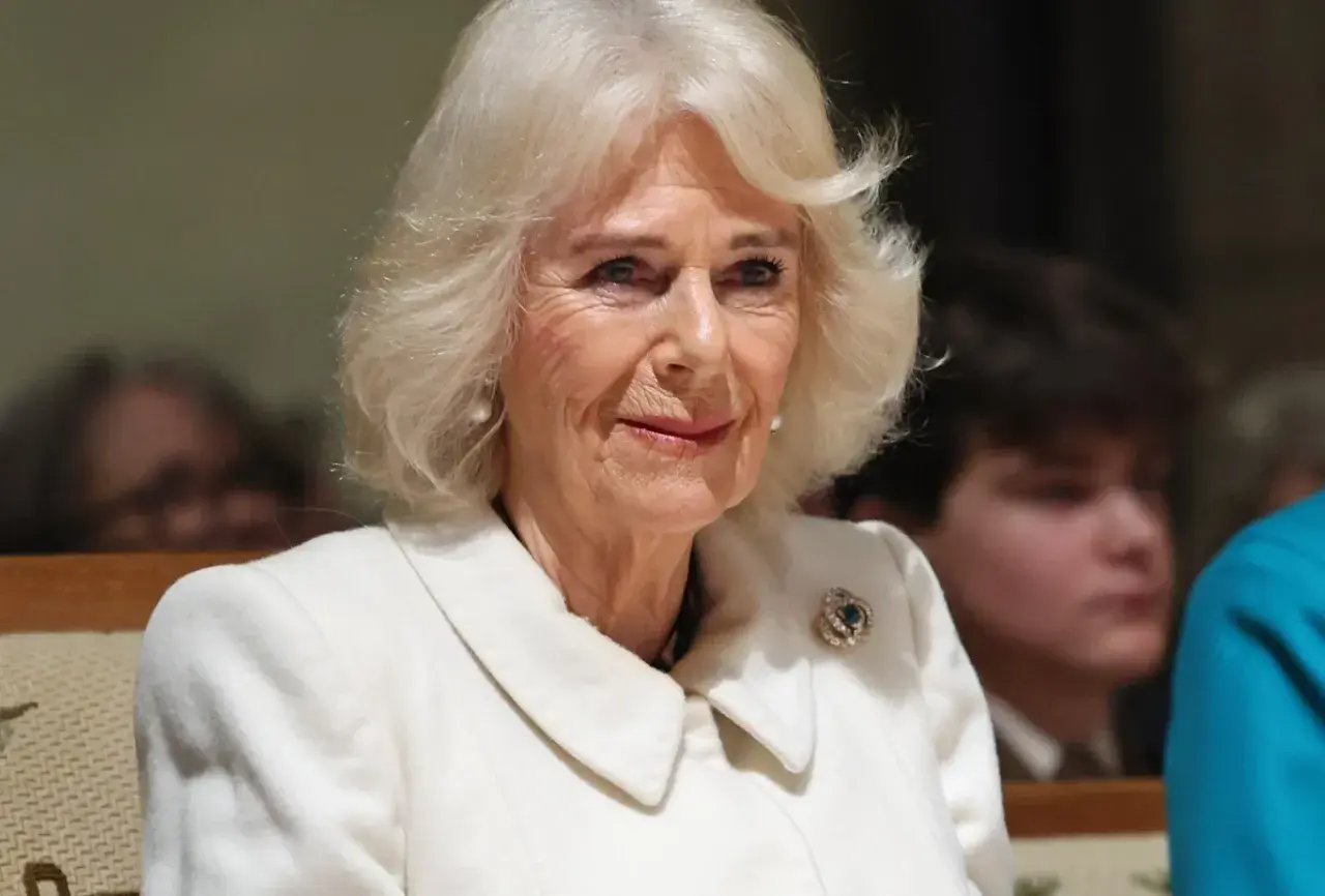 Uśmiechnięta królowa Camilla w białym płaszczu z broszką.