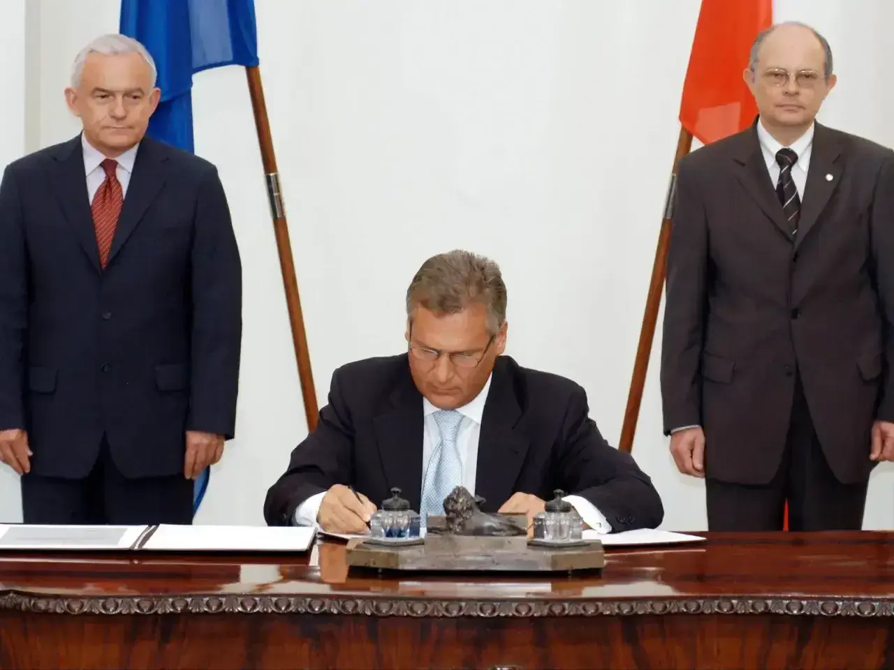 Uroczyste podpisanie dokumentów, kiedy Polska weszła do Unii. Trzech mężczyzn w garniturach, dwa flagi.