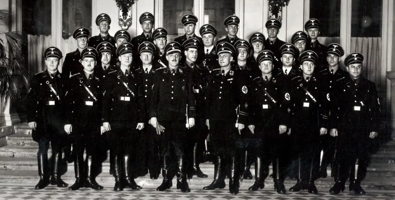 Grupa mężczyzn w czarnych mundurach i czapkach z orłem. Wśród nich widać symbole nawiązujące do gestapo.