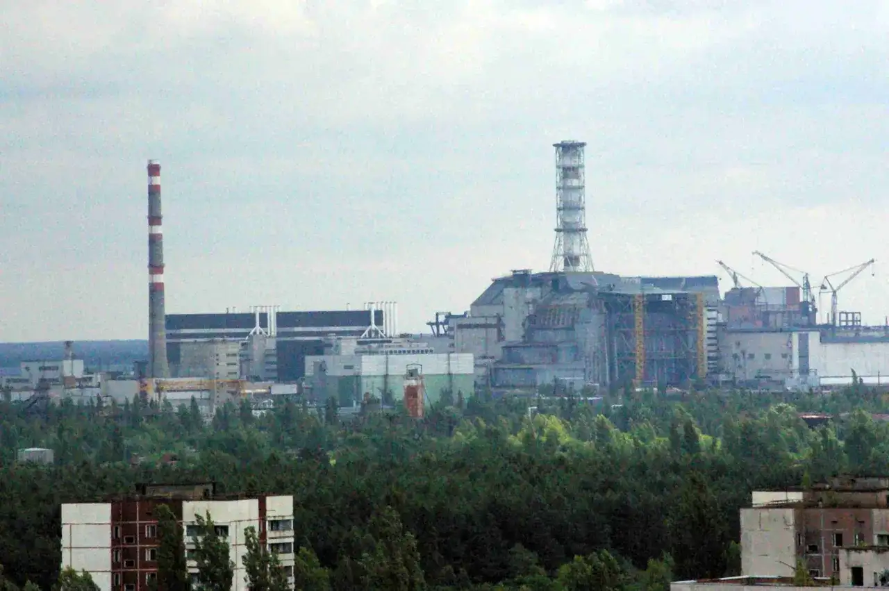 Widok na elektrownię w Czarnobylu, z charakterystycznym kominem i budynkami przemysłowymi otoczonymi lasem.