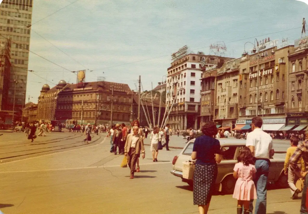 Tłoczny plac w Jugosławii, ludzie spacerują, jeżdżą tramwaje i samochody.