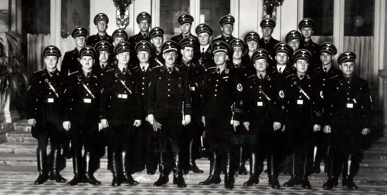 Grupa mężczyzn w czarnych mundurach i czapkach z orłem. Wśród nich widać symbole nawiązujące do gestapo.
