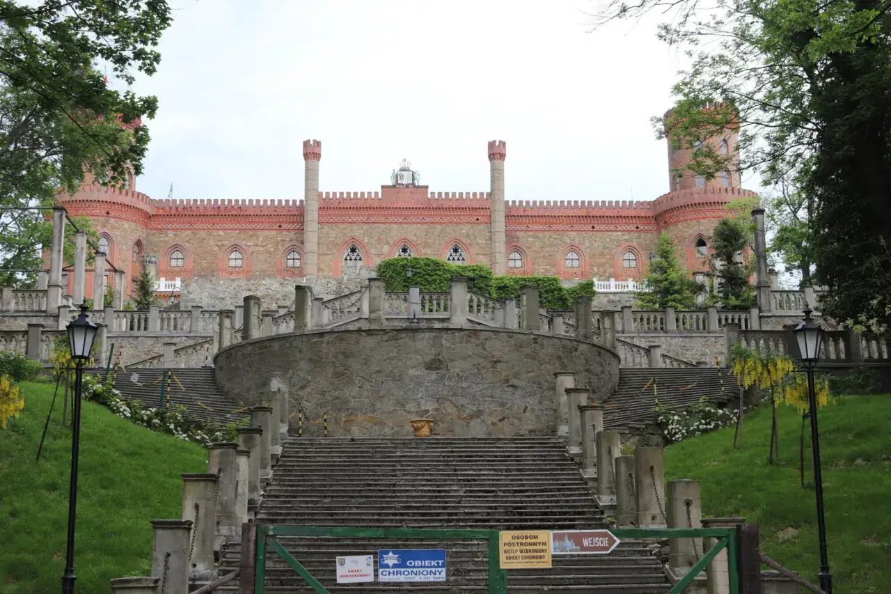 Kamieniec Ząbkowicki zamek, imponująca budowla z cegły i kamienia, otoczona zielenią, z monumentalnymi schodami prowadzącymi do wejścia.
