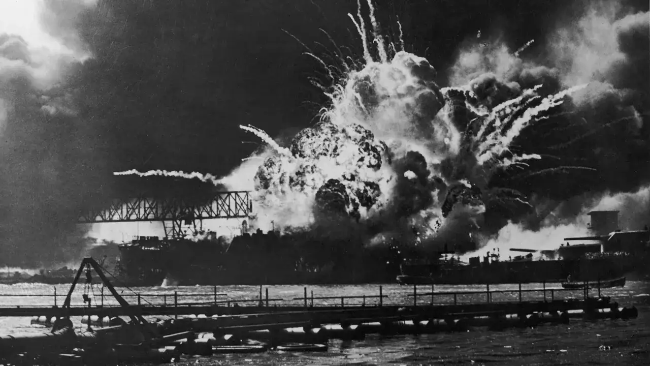 Potężna eksplozja w Pearl Harbor, płonące okręty i dym unoszący się nad wodą.
