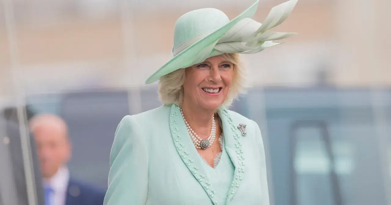 Uśmiechnięta królowa Camilla w miętowym kapeluszu i kostiumie, ozdobiona perłami i broszką.