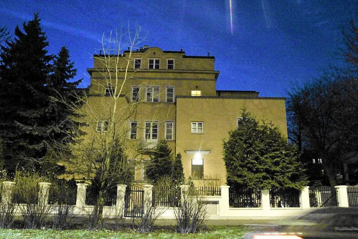 Żółty budynek nocą, otoczony drzewami i ogrodzeniem. W tle niebo z jasnym pasem, jakby ślad po meteorycie. Może to siedziba Opus Dei?