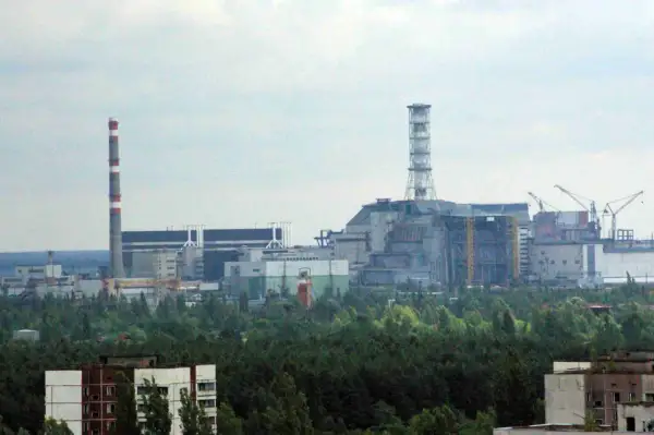 Widok na elektrownię w Czarnobylu, z charakterystycznym kominem i budynkami przemysłowymi otoczonymi lasem.