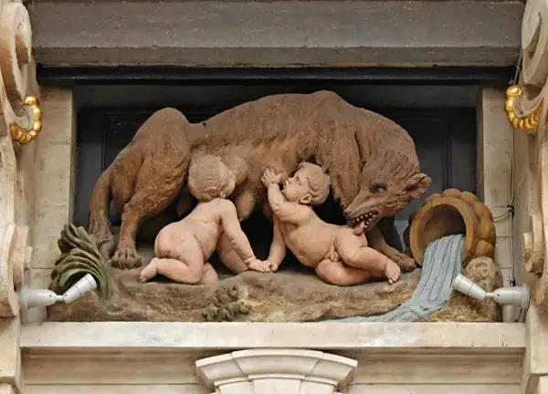 Rzeźba przedstawia wilczycę karmiącą Romulusa i Remusa, symbolizującą legendę o założeniu Rzymu. To klasyczny przykład motywu salut rzymski.