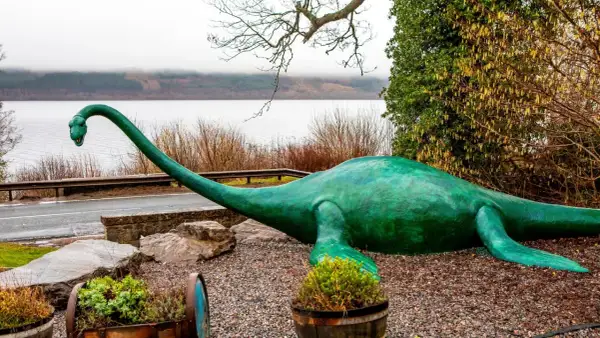 Zielony potwór z Loch Ness leży na kamieniach, z długą szyją wyciągniętą w stronę jeziora.