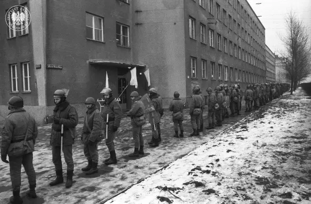 Zomo w zimowej scenerii, w pełnym rynsztunku, stoi w kolejce przed budynkiem.